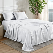 White satin bedding set on a bed in a bedroom with a plant and window in the background.