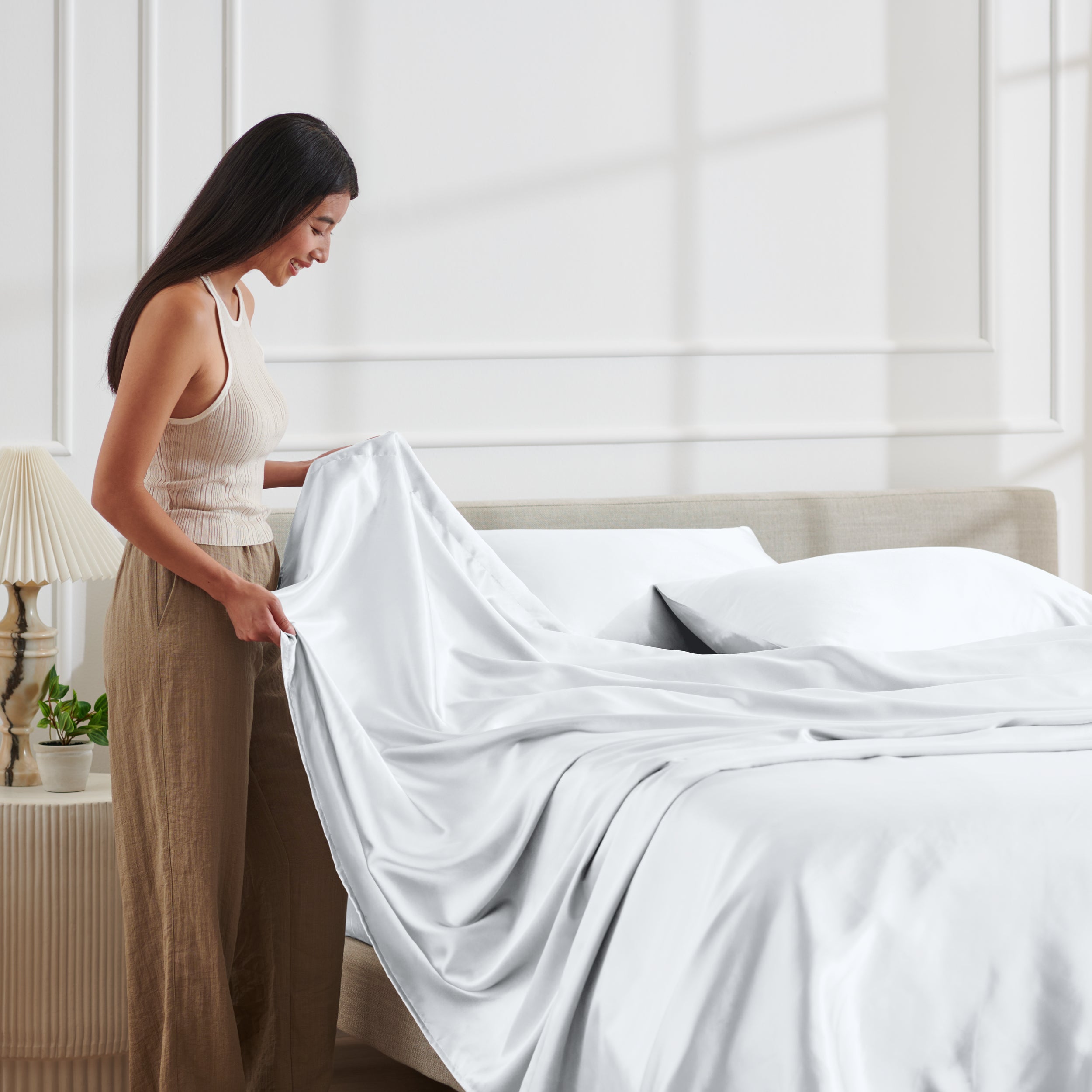 Woman arranging satin sheets on a bed in a bright bedroom.