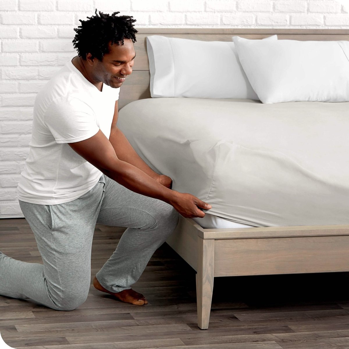 A man is kneeing and putting the corner of the fitted sheet on the mattress. The mattress is on a wooden bed frame. 