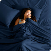 A smiling woman is lying in bed with a top sheet pulled up to her chin