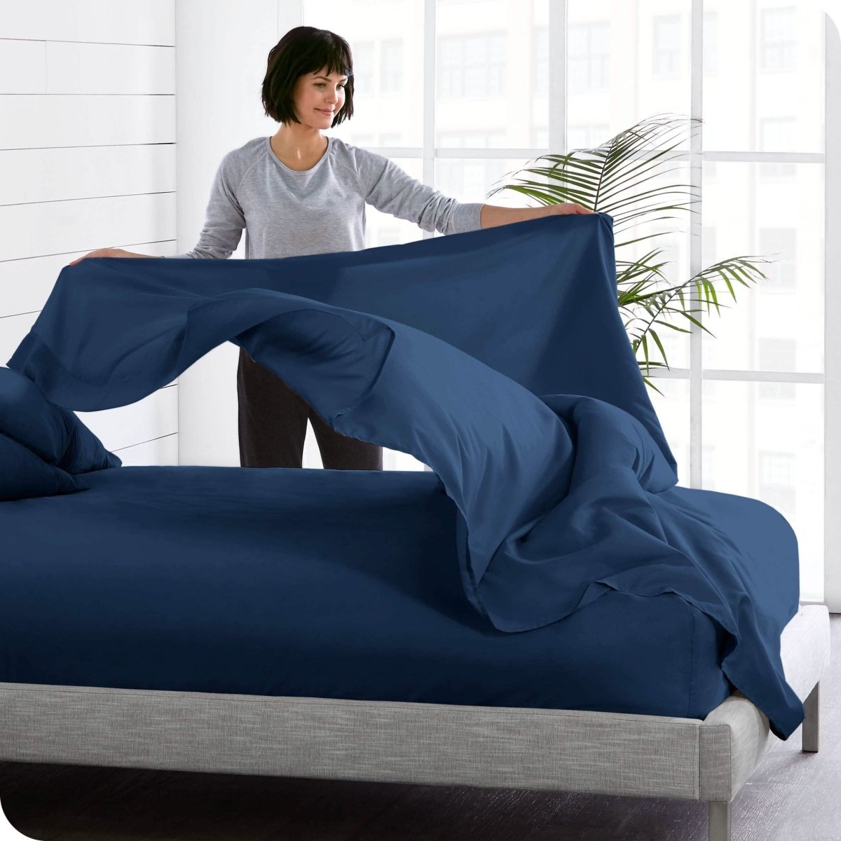 A woman is standing at the side of a bed putting a flat sheet on top of the mattress which has a fitted sheet on it