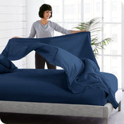 A woman is standing at the side of a bed putting a flat sheet on top of the mattress which has a fitted sheet on it