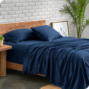 A side view of a modern bed with a microfiber sheet set on it. The bed has two pillows, a fitted sheet, and a flat sheet on the mattress. The bed is placed next to a side table.