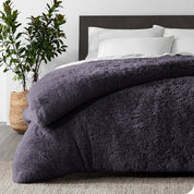 A eggplant shaggy duvet cover on a bed with white sheets and pillowcases. A large plant is next to the bed.
