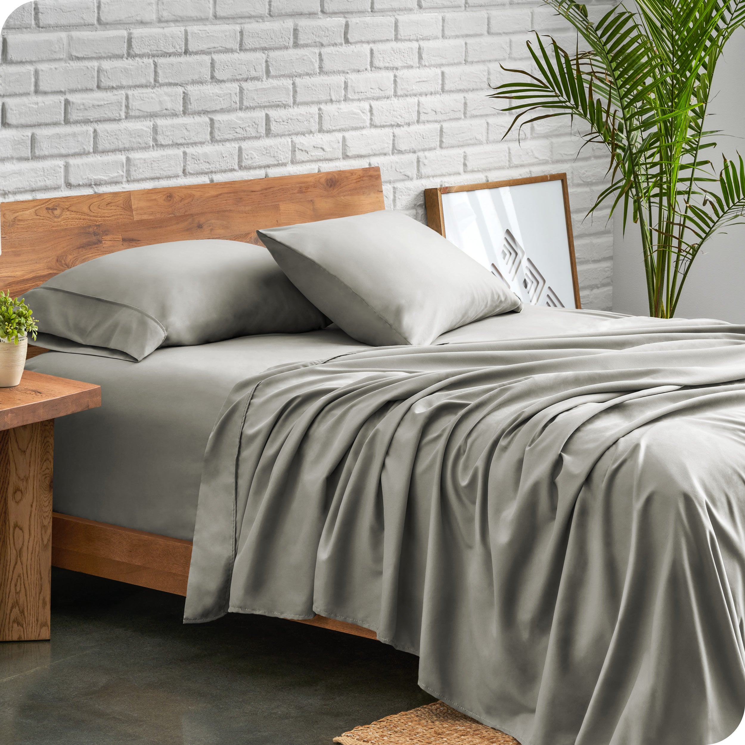 A side view of a modern bed with a microfiber sheet set on it. The bed has two pillows, a fitted sheet, and a flat sheet on the mattress. The bed is placed next to a side table.