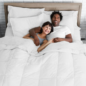 A happy couple (male and female) lying on the modern bed under a white comforter and a few pillows.