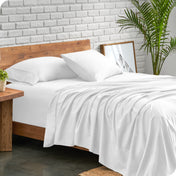 A side view of a modern bed with a microfiber sheet set on it. The bed has two pillows, a fitted sheet, and a flat sheet on the mattress. The bed is placed next to a side table.