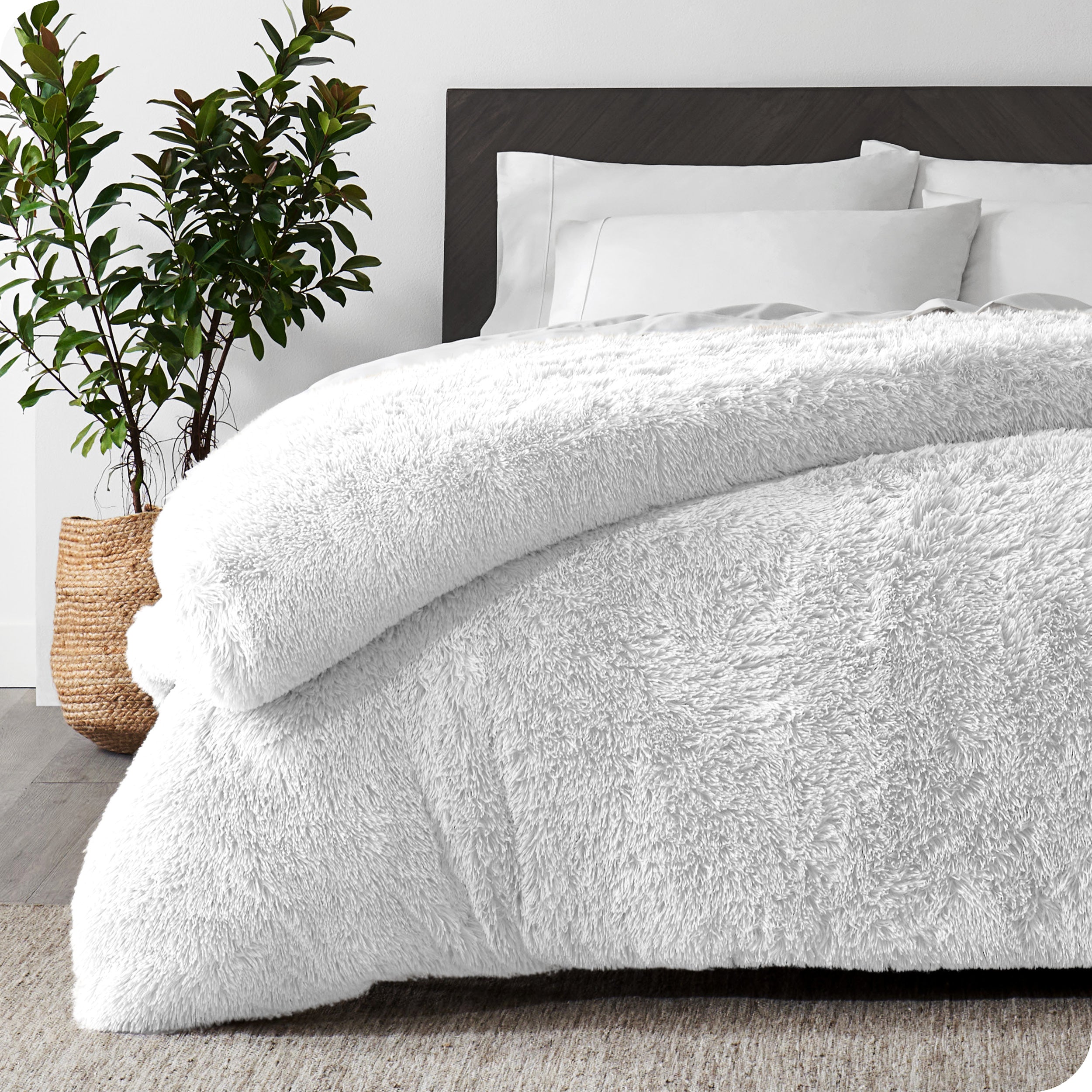 A white shaggy duvet cover on a bed with white sheets and pillowcases. A large plant is next to the bed.