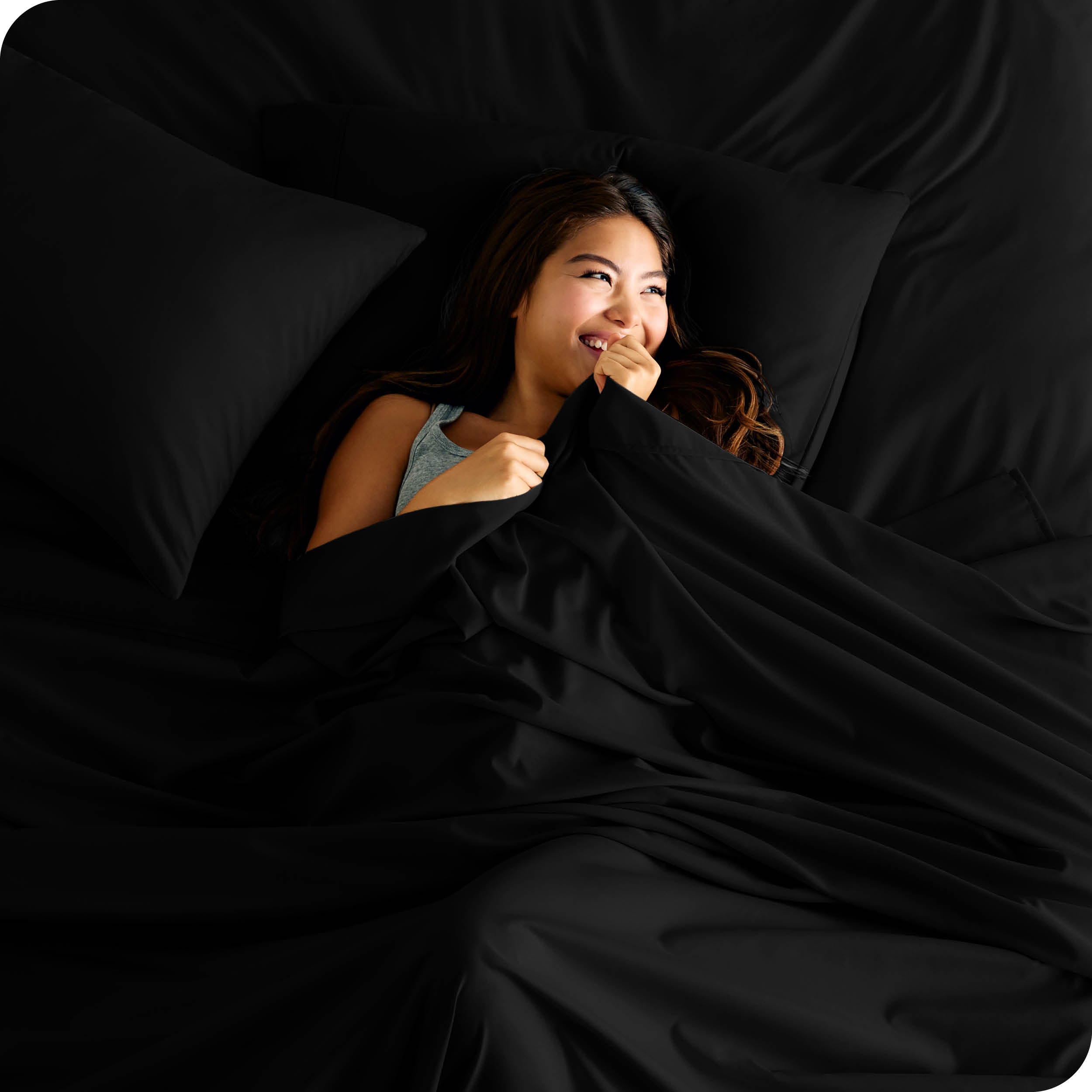 A smiling woman is lying in bed with a top sheet pulled up to her chin