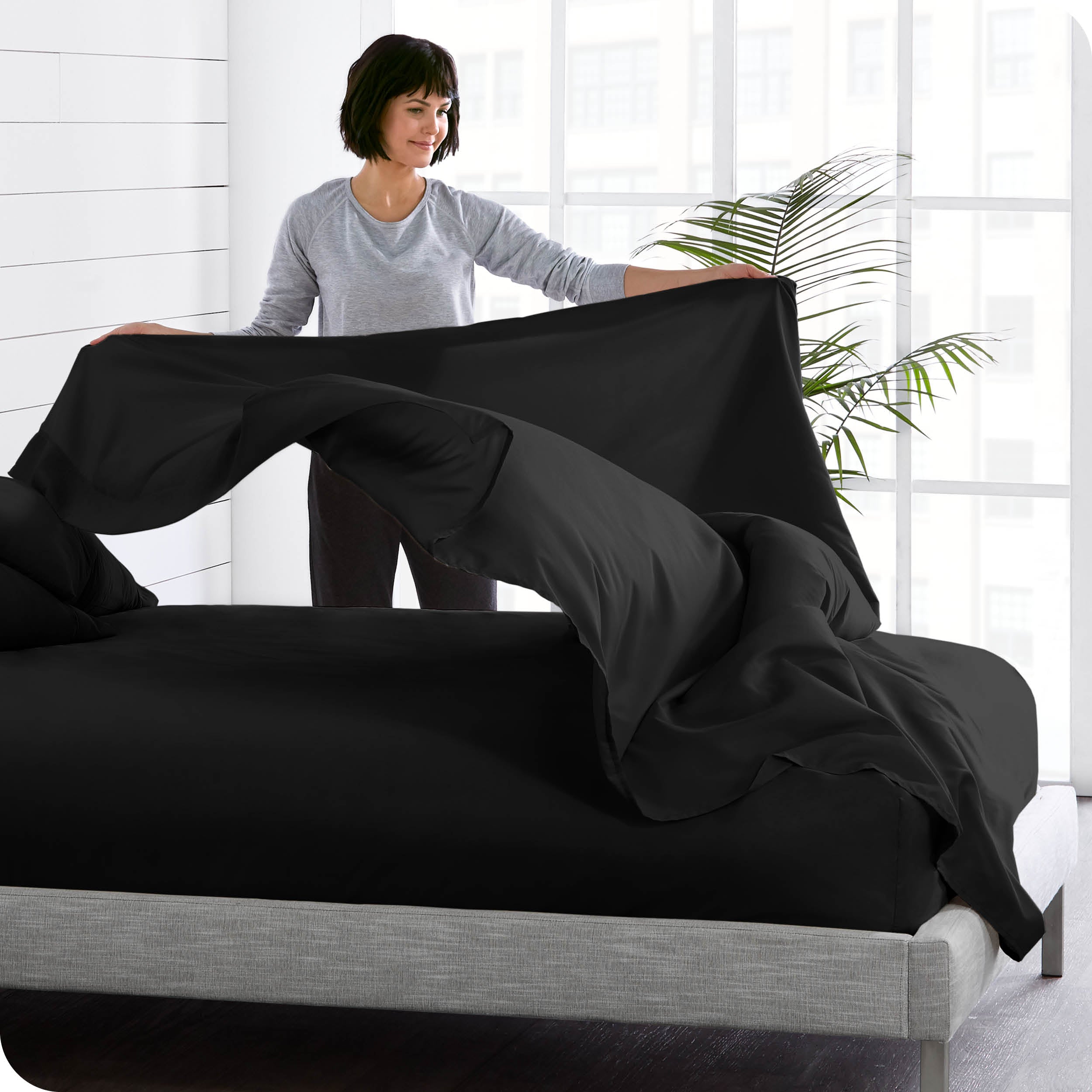 A woman is standing at the side of a bed putting a flat sheet on top of the mattress which has a fitted sheet on it