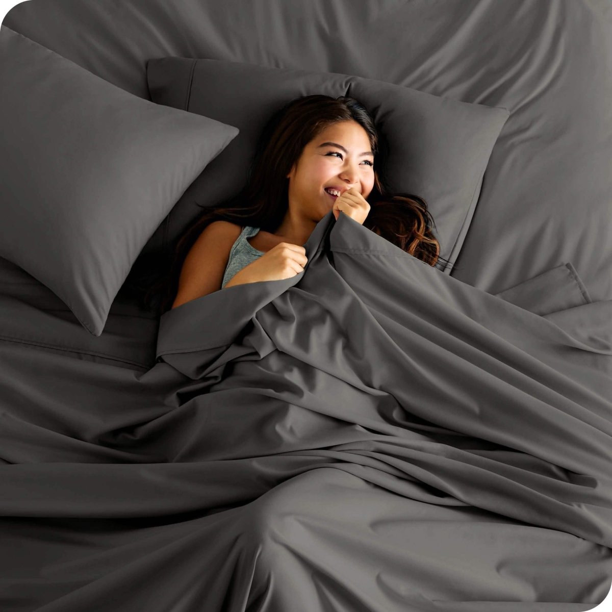 A smiling woman is lying in bed with a top sheet pulled up to her chin
