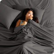 A smiling woman is lying in bed with a top sheet pulled up to her chin