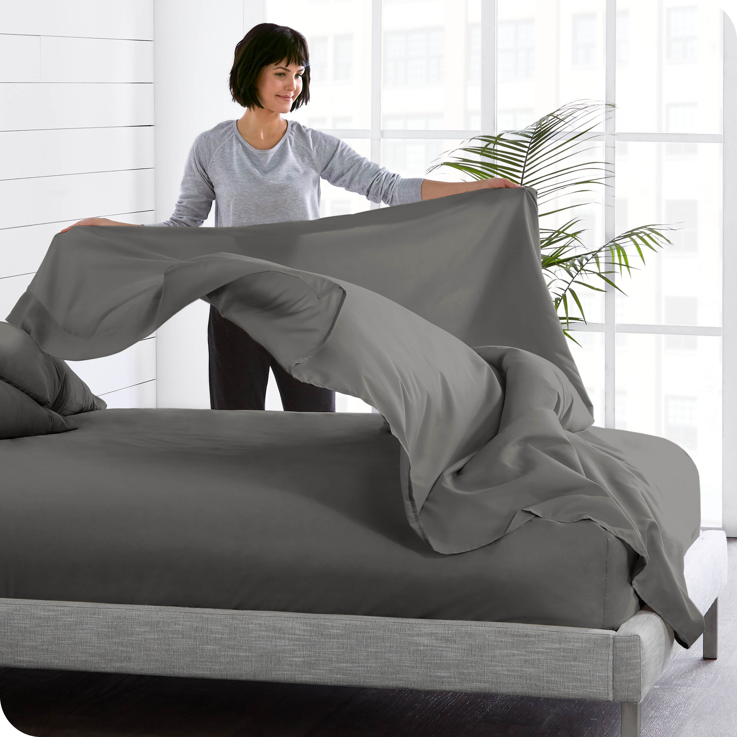 A woman is standing at the side of a bed putting a flat sheet on top of the mattress which has a fitted sheet on it