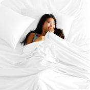 A smiling woman is lying in bed with a top sheet pulled up to her chin