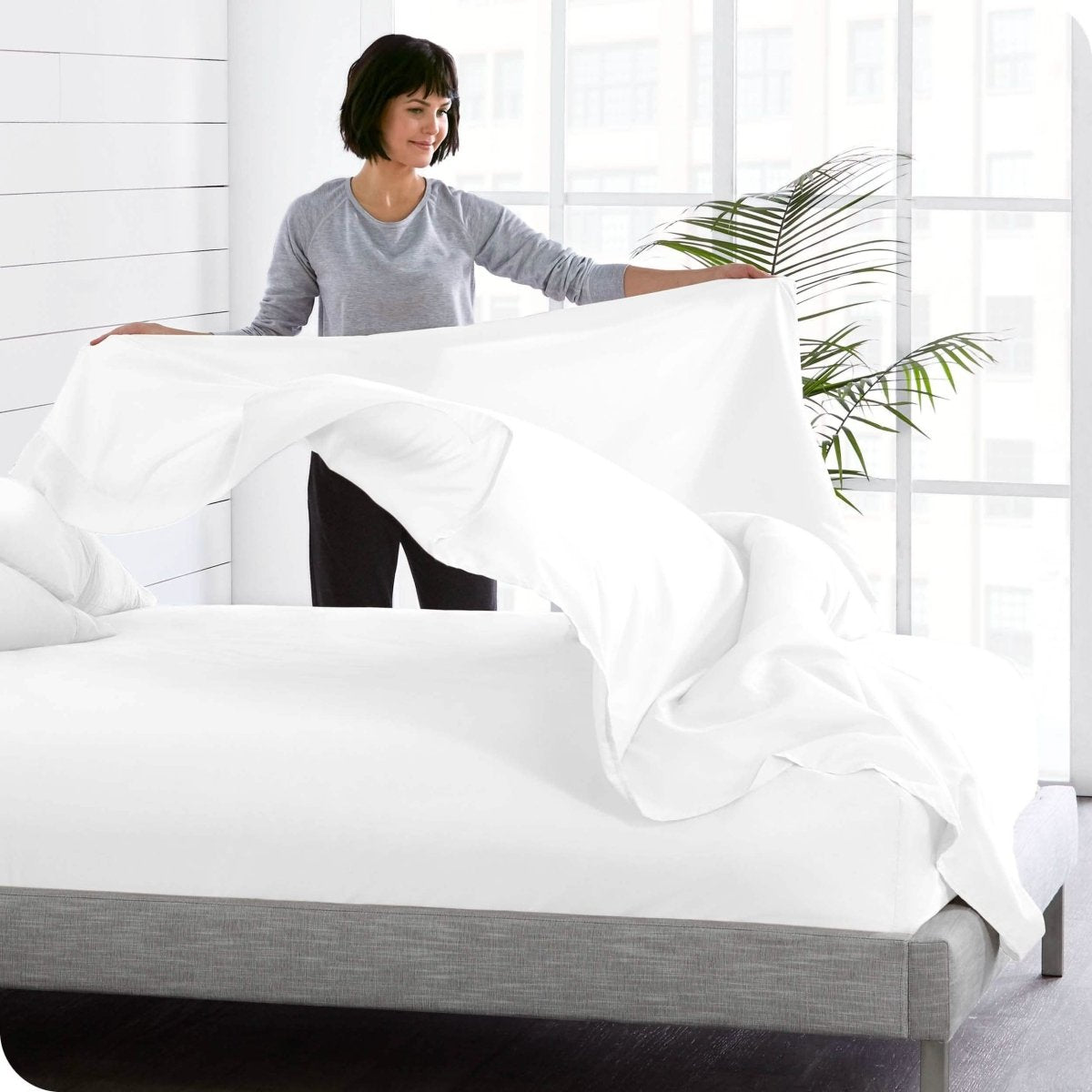 A woman is standing at the side of a bed putting a flat sheet on top of the mattress which has a fitted sheet on it