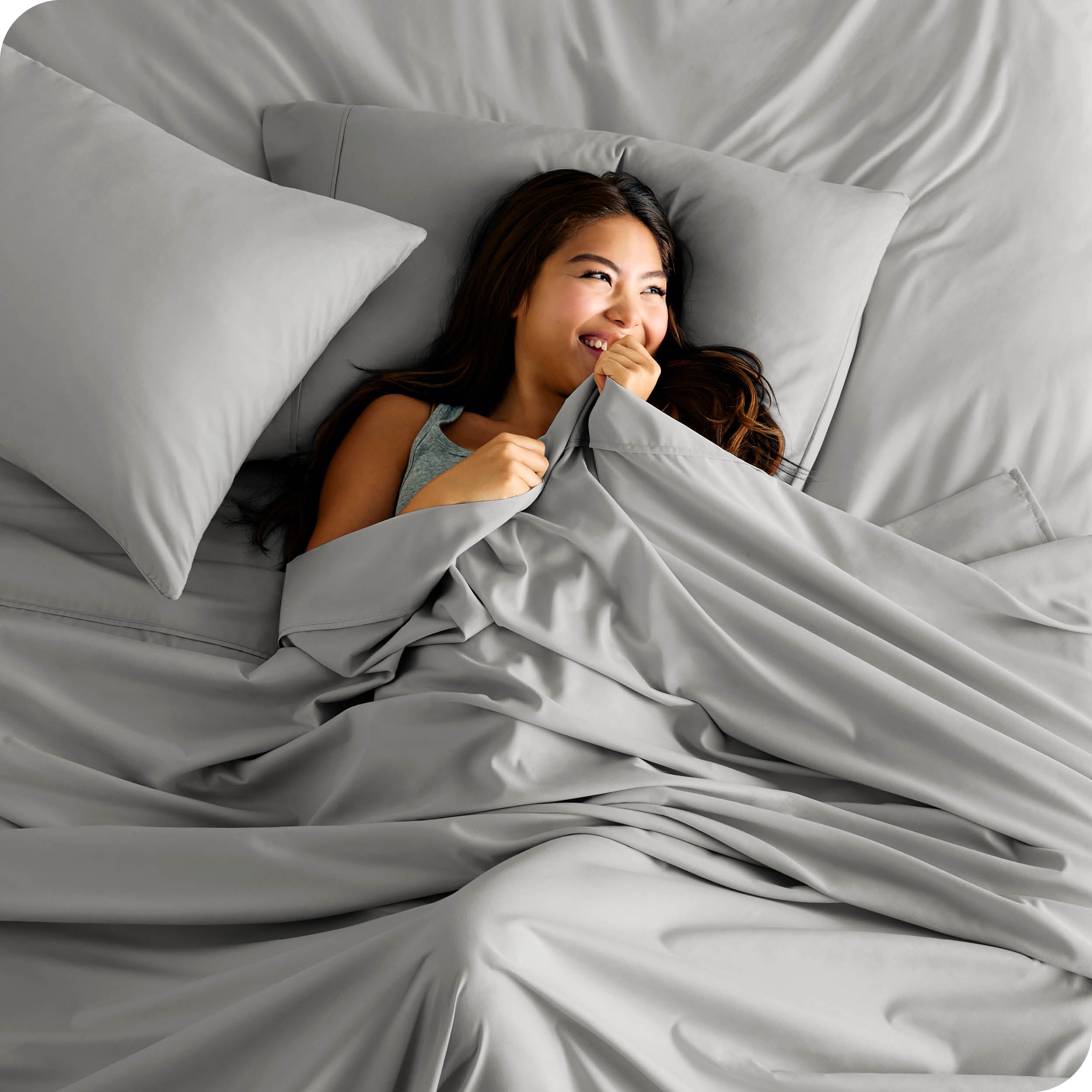 A smiling woman is lying in bed with a top sheet pulled up to her chin