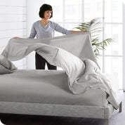A woman is standing at the side of a bed putting a flat sheet on top of the mattress which has a fitted sheet on it